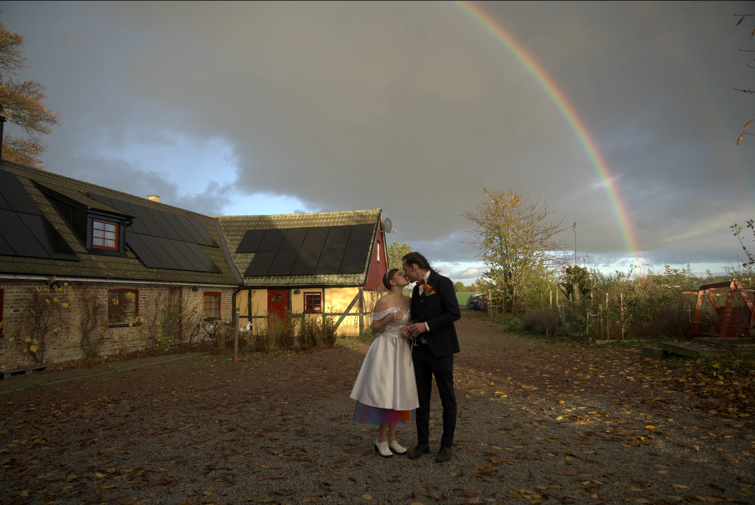 Brudpar kysser varandra utomhus med en regnbåge i bakgrunden.